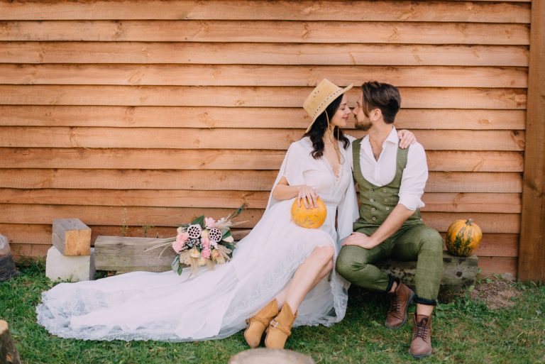 novomanželé oblečené v boho stylu sedí u dřevěné stěny s dýní